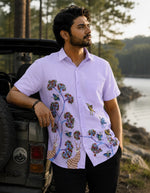 Load image into Gallery viewer, Gond hand-painted linen shirt featuring birds, branching tree forms, and deer motifs flowing across the front panel, observed while leaning against an open jeep beside a quiet lake and watching the surrounding trees within a soft golden-hour wilderness setting.