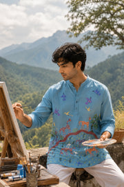 Artist painting at an easel while wearing the Bhil Pithora Horses hand-painted 100% linen short kurta, exploring the relationship between clothing, art, and creative expression in a natural outdoor setting.