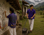 Bhil hand-painted linen shirt featuring scattered botanical motifs across the front and a large Mahua tree composition across the back with male and female figures alongside a child gathering fallen Mahua blossoms beneath branching forms, accompanied by grazing deer and birds among fruit-bearing branches, photographed in a quiet rural village setting while painting mural motifs on a traditional house wall and maintaining the surrounding farm landscape.