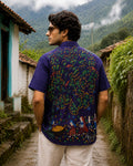 Bhil hand-painted linen shirt featuring a large Mahua tree artwork across the back with male and female figures alongside a child collecting fallen Mahua blossoms near the trunk, birds among fruit-bearing branches, grazing deer, and scattered Mahua blossoms and fruits, photographed while walking through a mist-covered village lane surrounded by monsoon greenery and stone pathways.