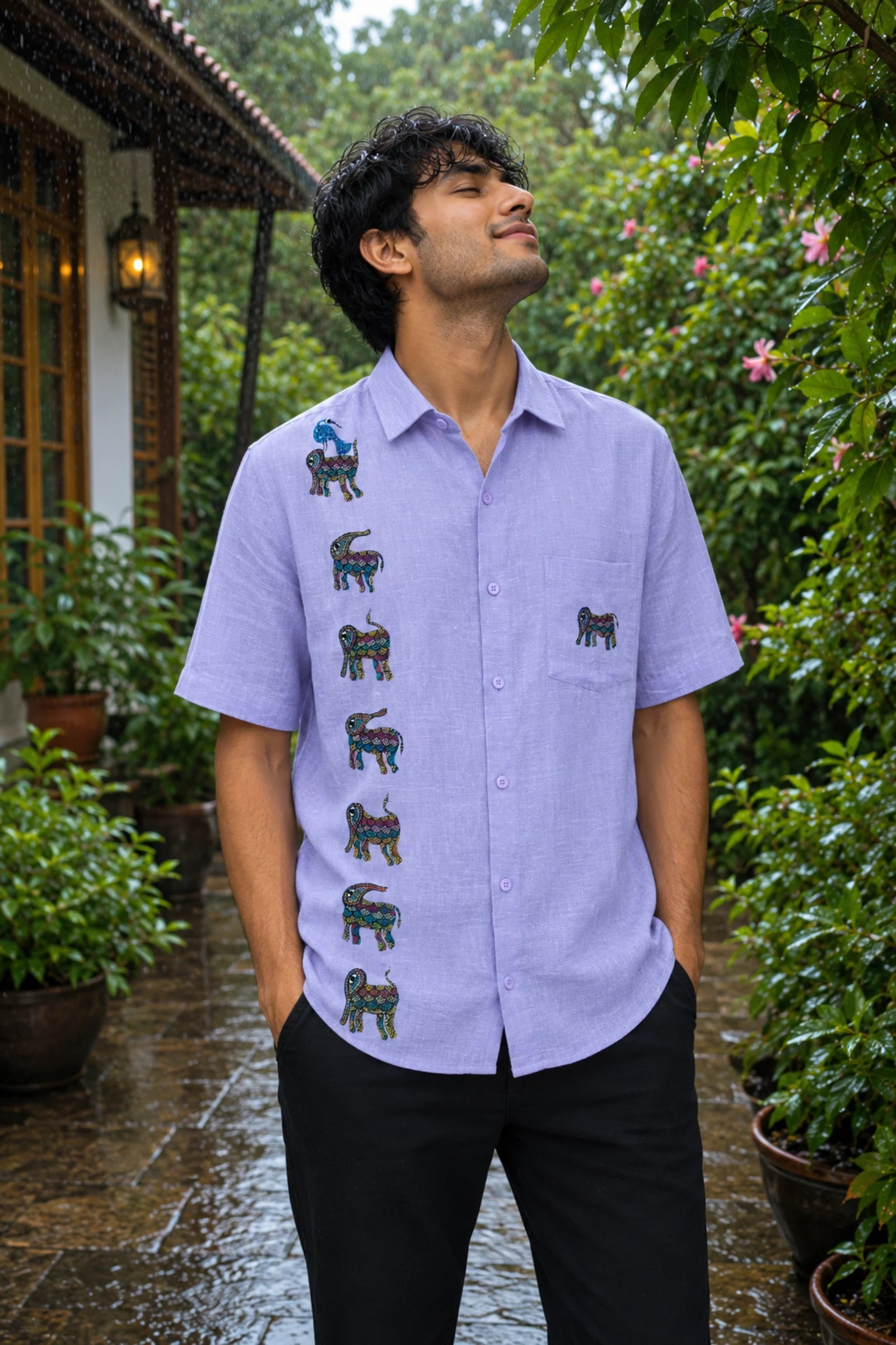 Bhil hand-painted linen shirt featuring seven elephant motifs arranged vertically across the front panel, a blue bird perched on the uppermost elephant, and an additional elephant motif on the pocket, photographed during a gentle monsoon drizzle within a private garden setting surrounded by flowering greenery and rain-soaked stone pathways.