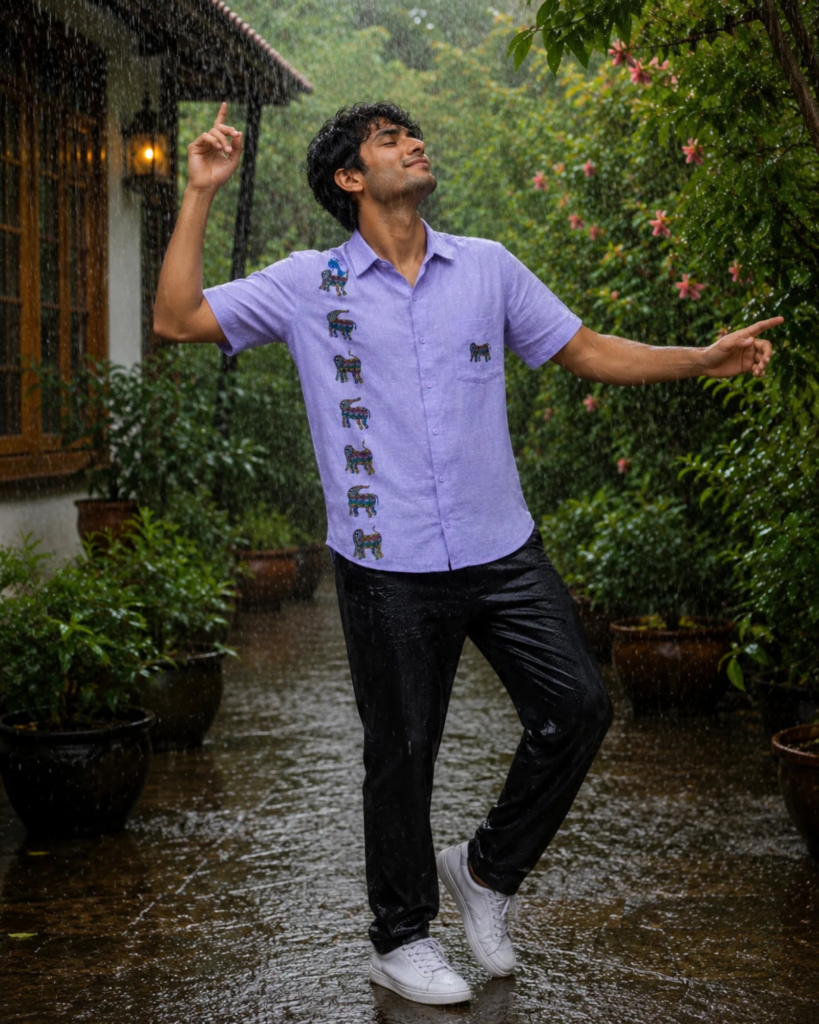 Bhil hand-painted lavender linen shirt featuring seven elephant motifs arranged vertically across the front panel, a blue bird perched on the uppermost elephant, and an additional elephant motif on the pocket, captured during a joyful monsoon rain dance within a lush garden courtyard, with wet stone pathways, flowering greenery, and falling rain creating a vibrant seasonal atmosphere.