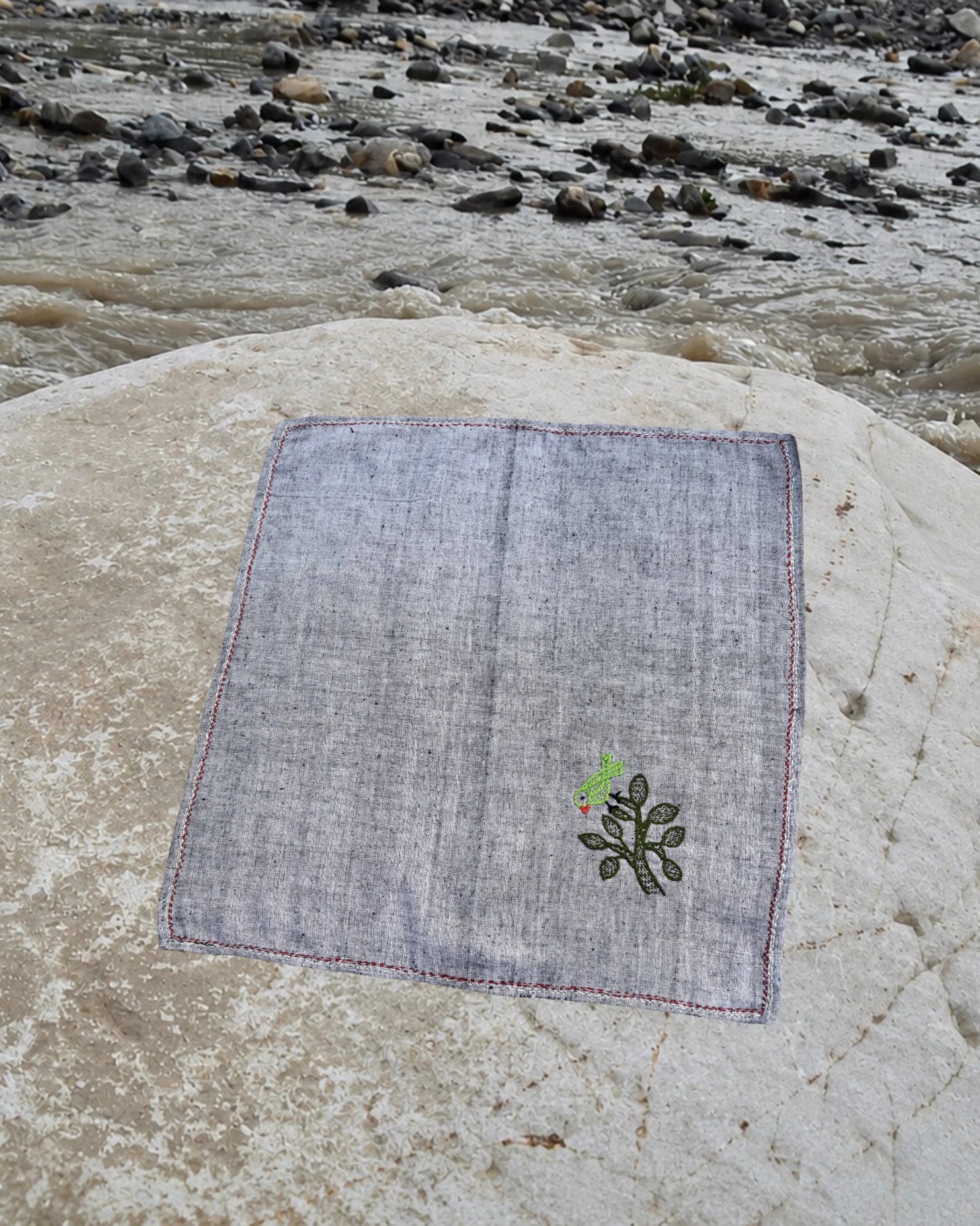 Hand-embroidered Bihar Sujani khadi handkerchief with a bird perched on a leafy tree artwork, photographed on a weathered river stone.