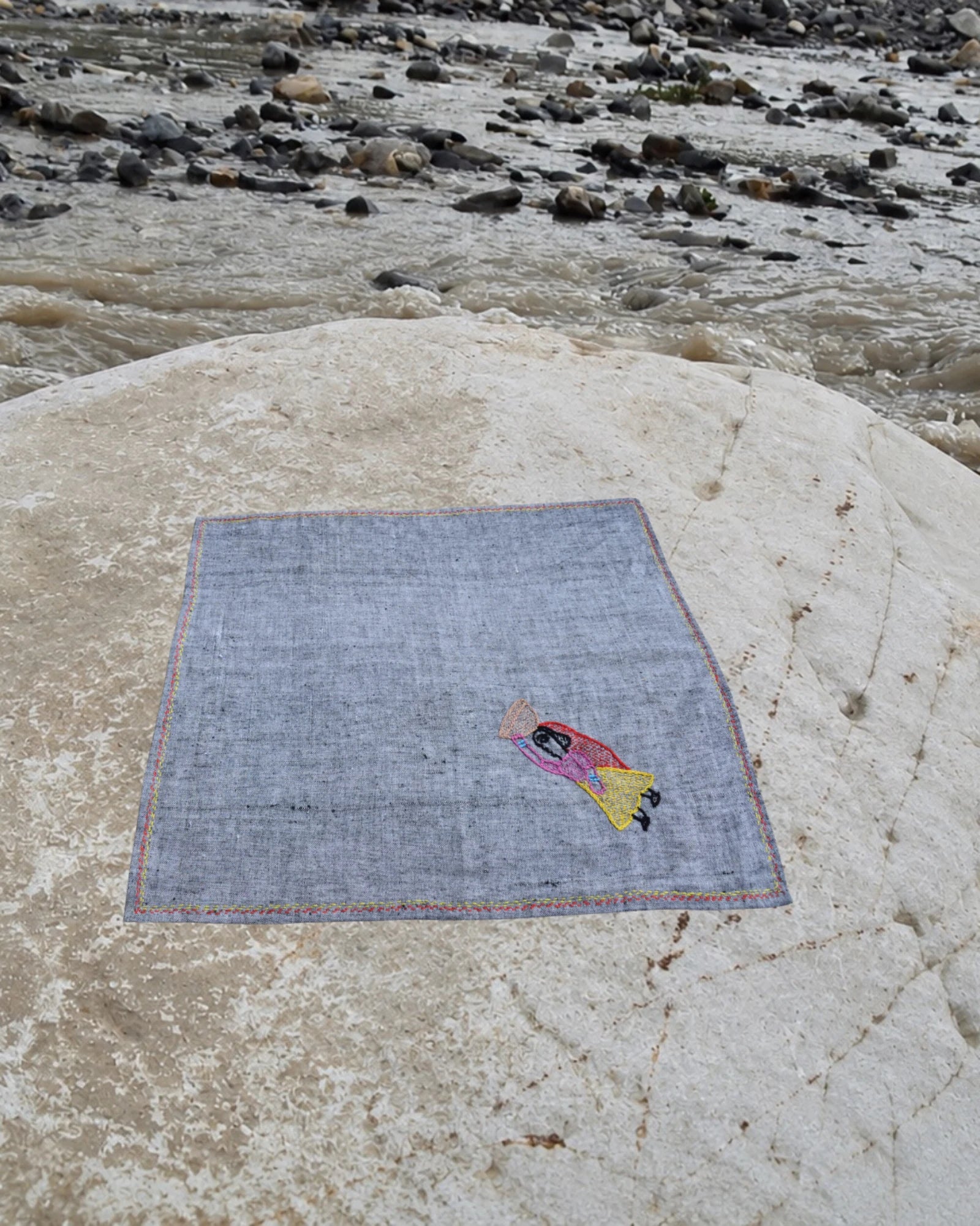 Hand-embroidered Bihar Sujani khadi handkerchief featuring a woman carrying a basket of food artwork, photographed on a weathered river stone.