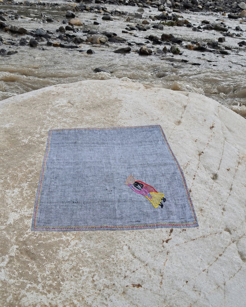 Hand-embroidered Bihar Sujani khadi handkerchief featuring a woman carrying a basket of food artwork, photographed on a weathered river stone.