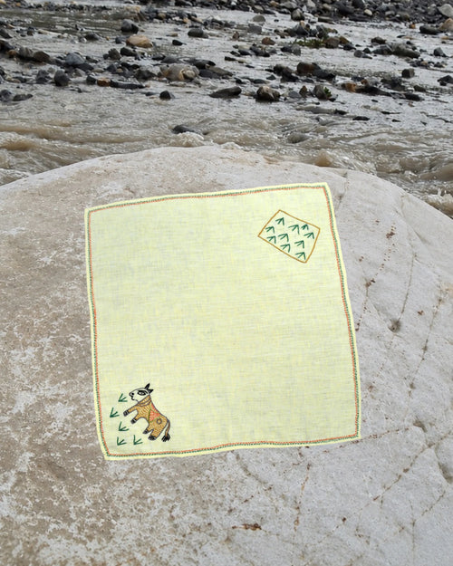 Hand-embroidered Bihar Sujani linen handkerchief featuring artwork of a cow grazing on grass in one corner and a cultivated field in the opposite corner, photographed on a weathered river stone.