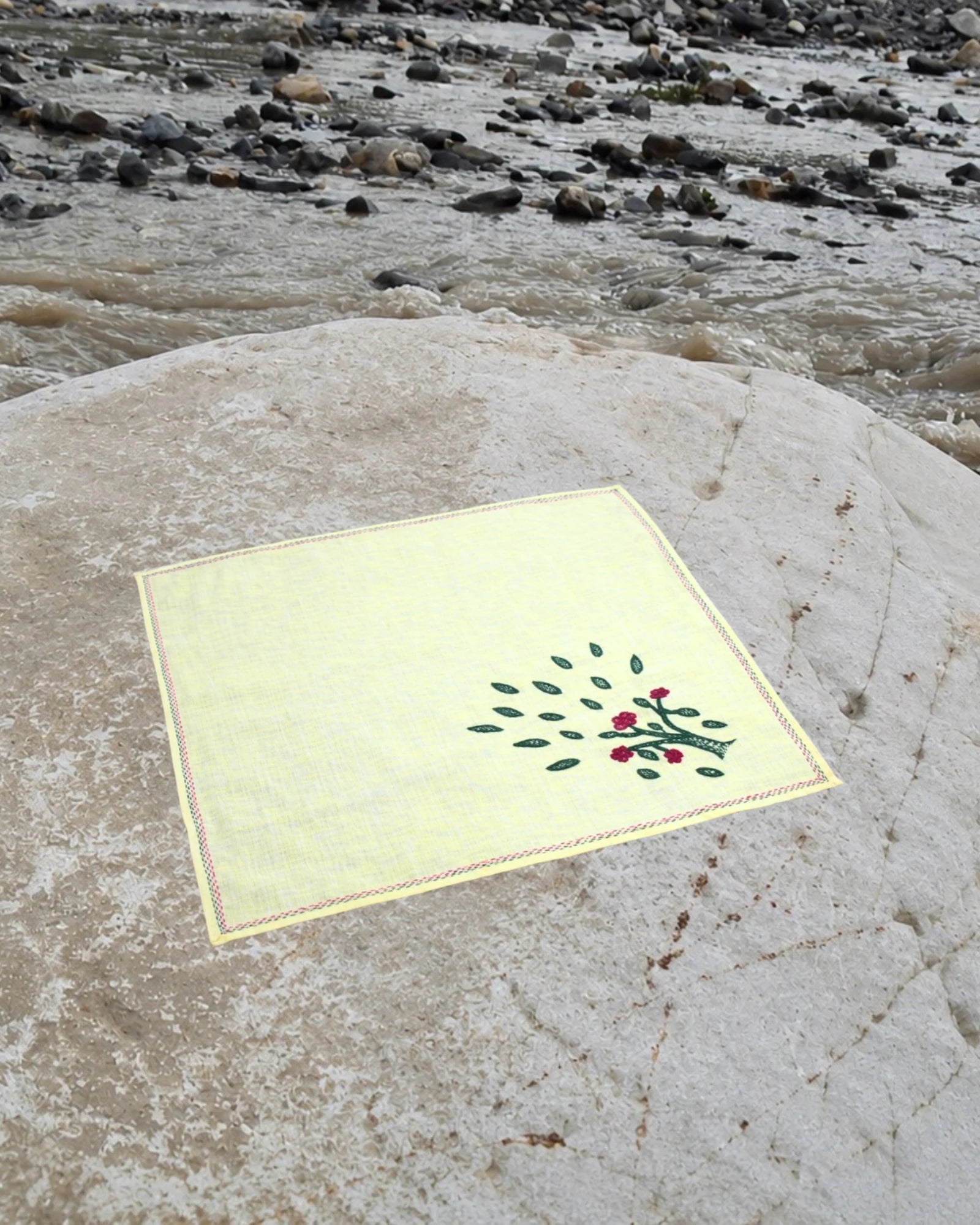 Bihar Sujani hand-embroidered linen handkerchief featuring a flowering tree artwork, photographed on a weathered river stone in a natural riverside setting.