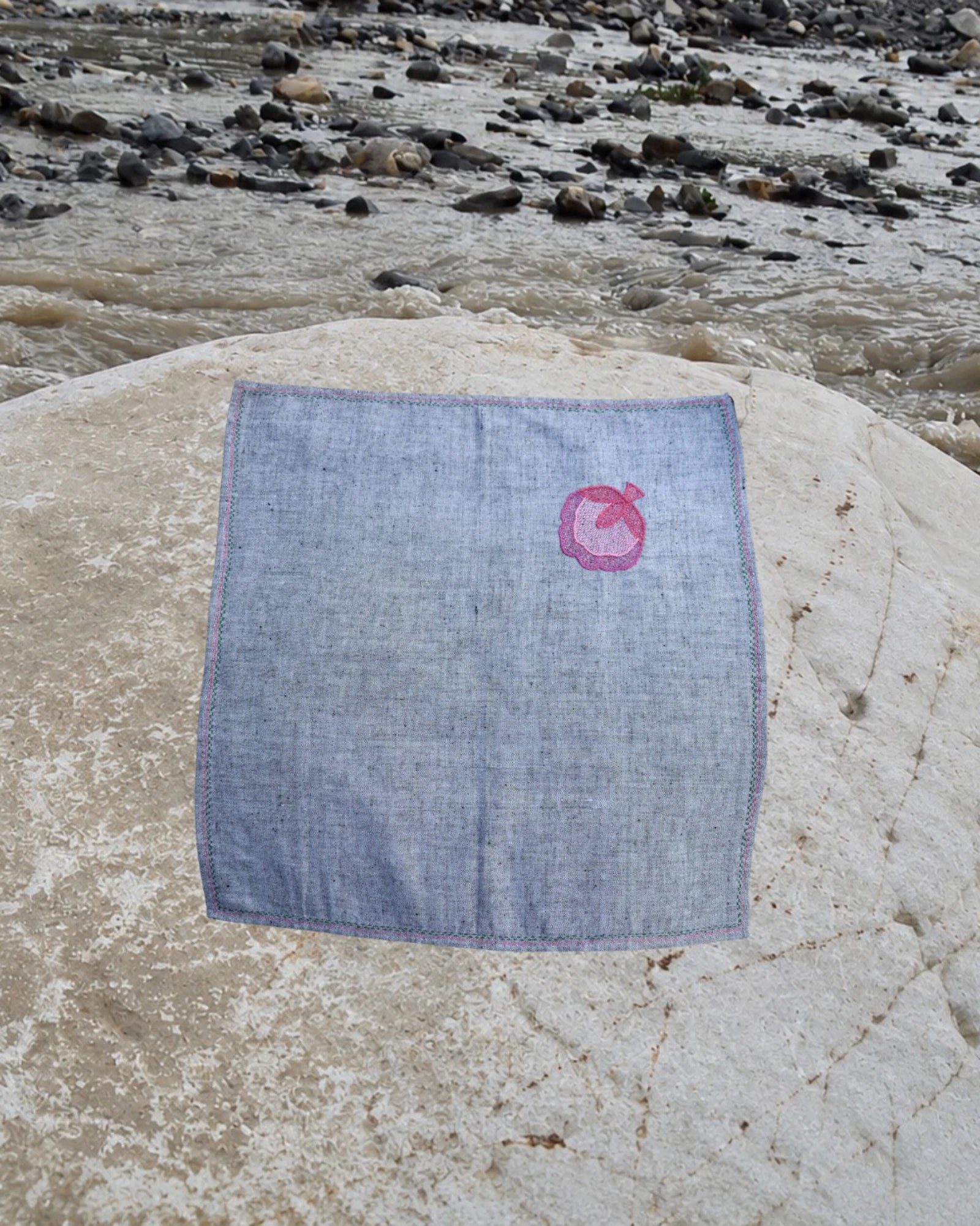 Hand-embroidered Bihar Sujani khadi cotton handkerchief featuring a pink flower blossom artwork in one corner, photographed on a weathered river stone.