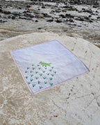 Hand-embroidered Bihar Sujani khadi cotton handkerchief featuring a green parrot artwork, displayed on a weathered river stone beside flowing water.