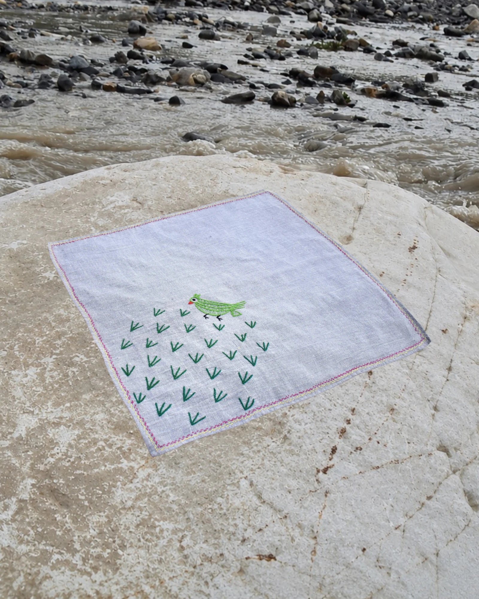 Hand-embroidered Bihar Sujani khadi cotton handkerchief featuring a green parrot artwork, displayed on a weathered river stone beside flowing water.