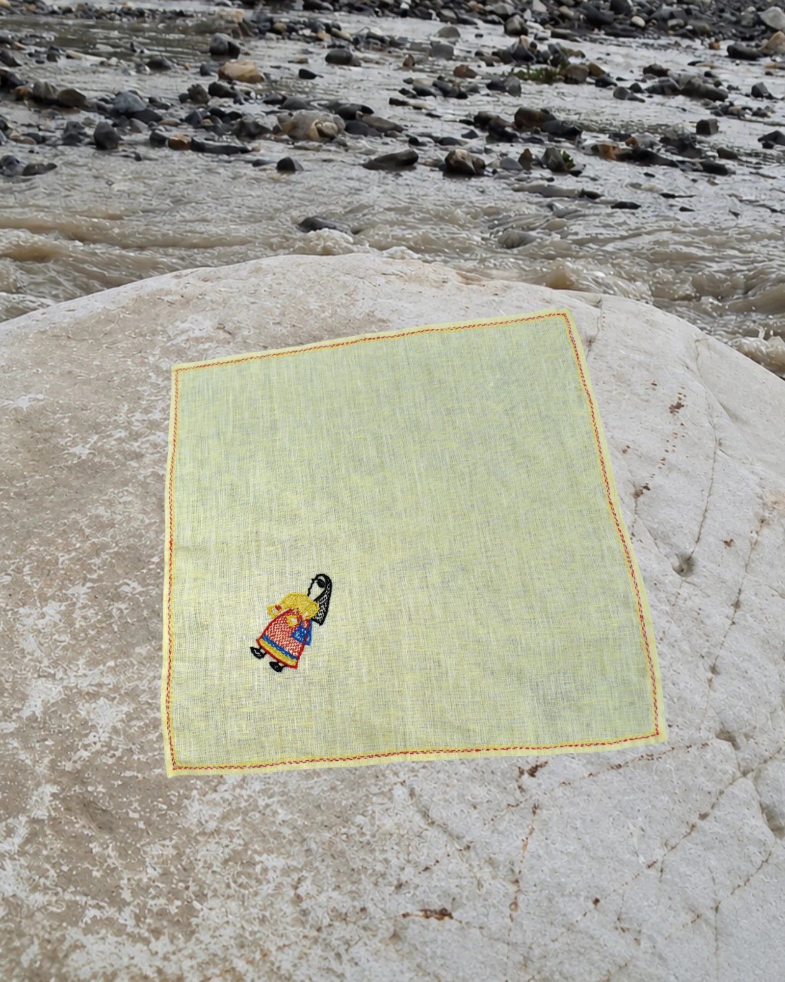 Hand-embroidered Bihar Sujani linen handkerchief featuring artwork of a woman carrying a shopping bag, photographed on a weathered river stone.