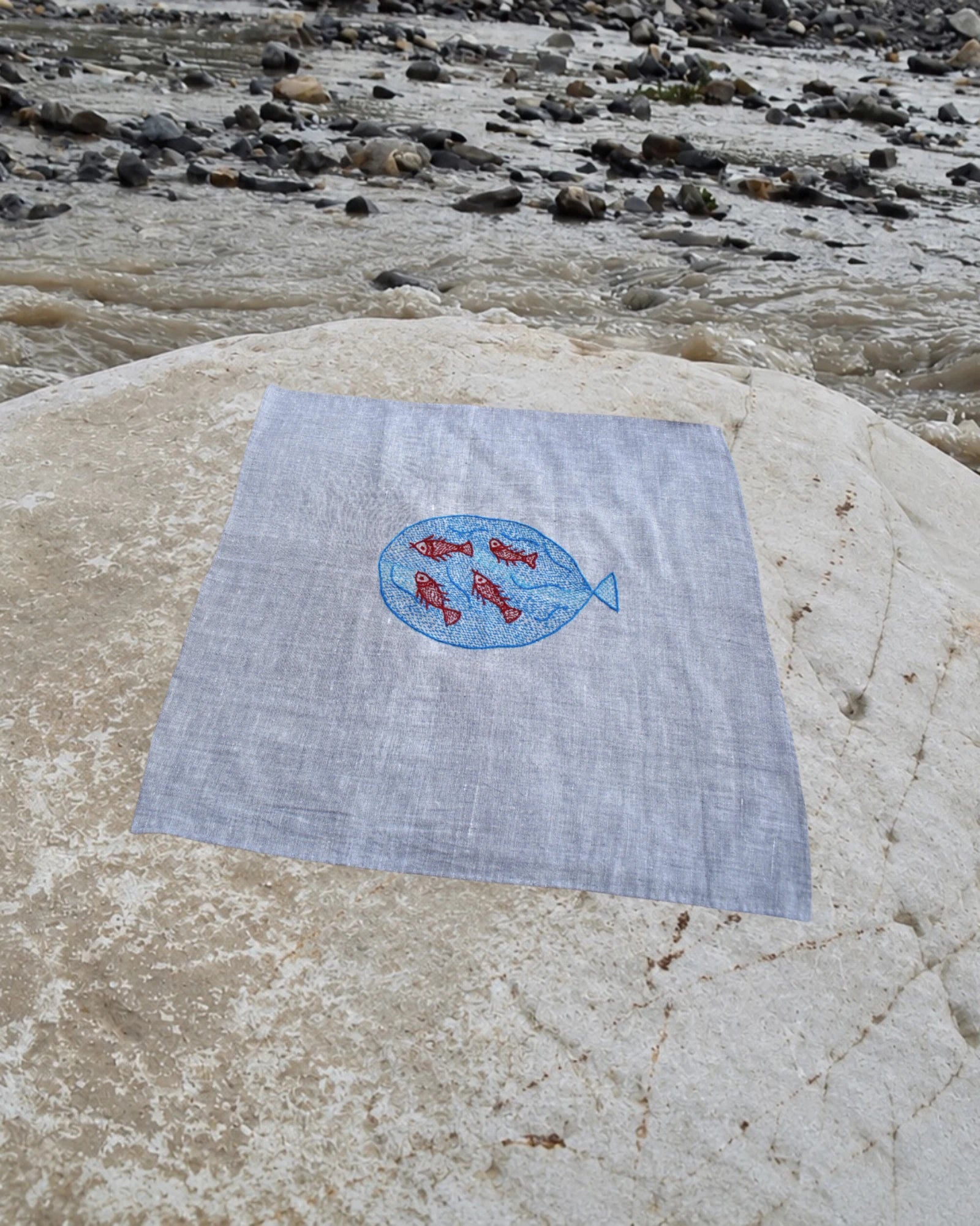 Hand-embroidered Bihar Sujani khadi handkerchief featuring a large blue fish artwork enclosing four red fish artworks, photographed on a weathered river stone.