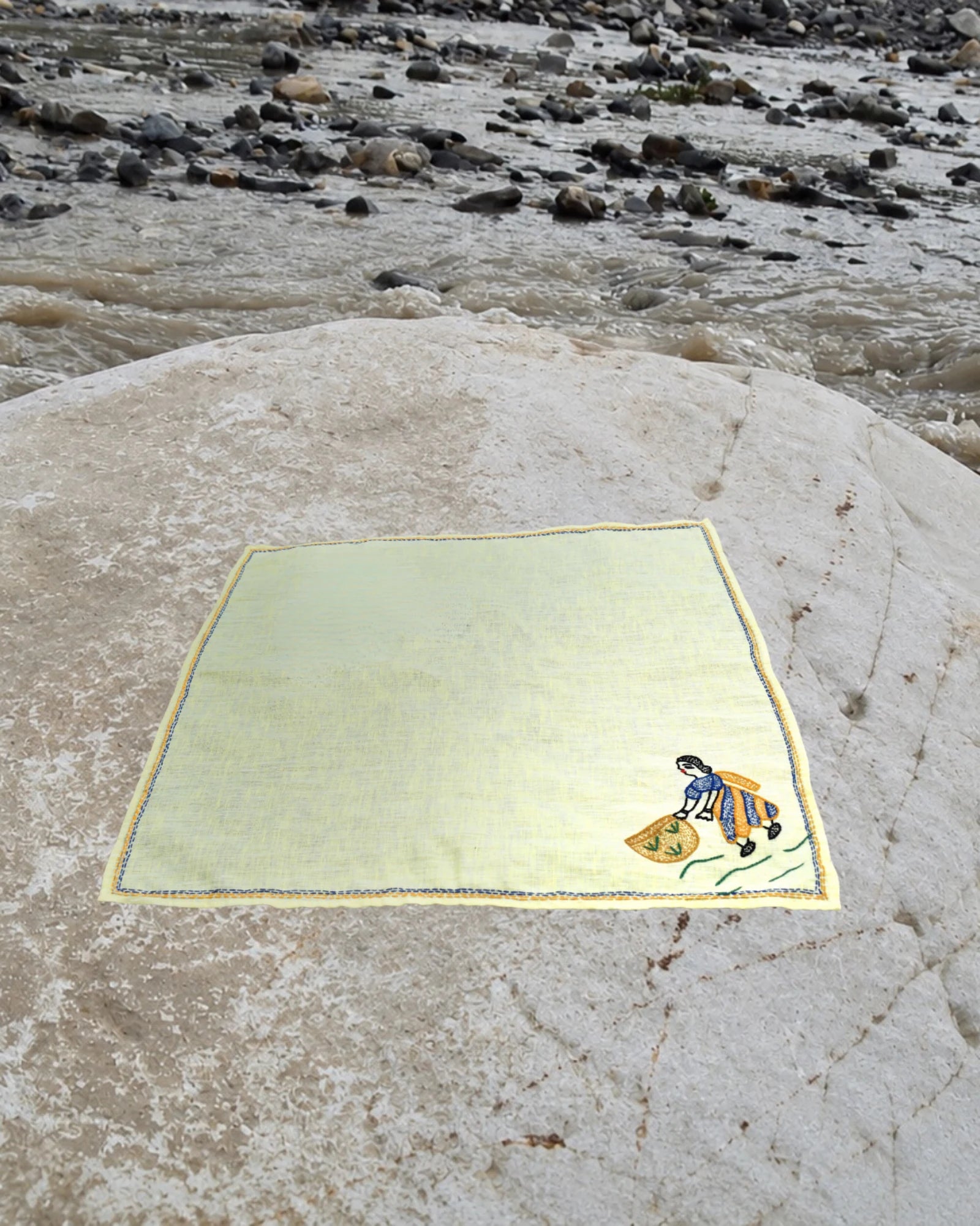 Bihar Sujani hand-embroidered linen handkerchief featuring artwork of a woman gathering the harvest, displayed on a weathered river stone beside a flowing river.