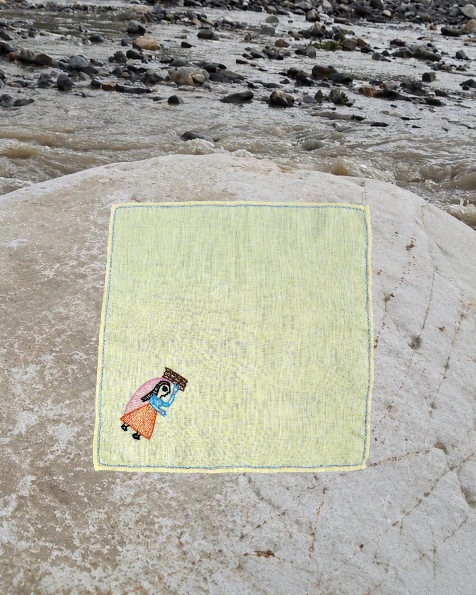 Hand-embroidered Bihar Sujani linen handkerchief featuring artwork of a woman carrying a bundle of firewood, photographed on a weathered river stone.