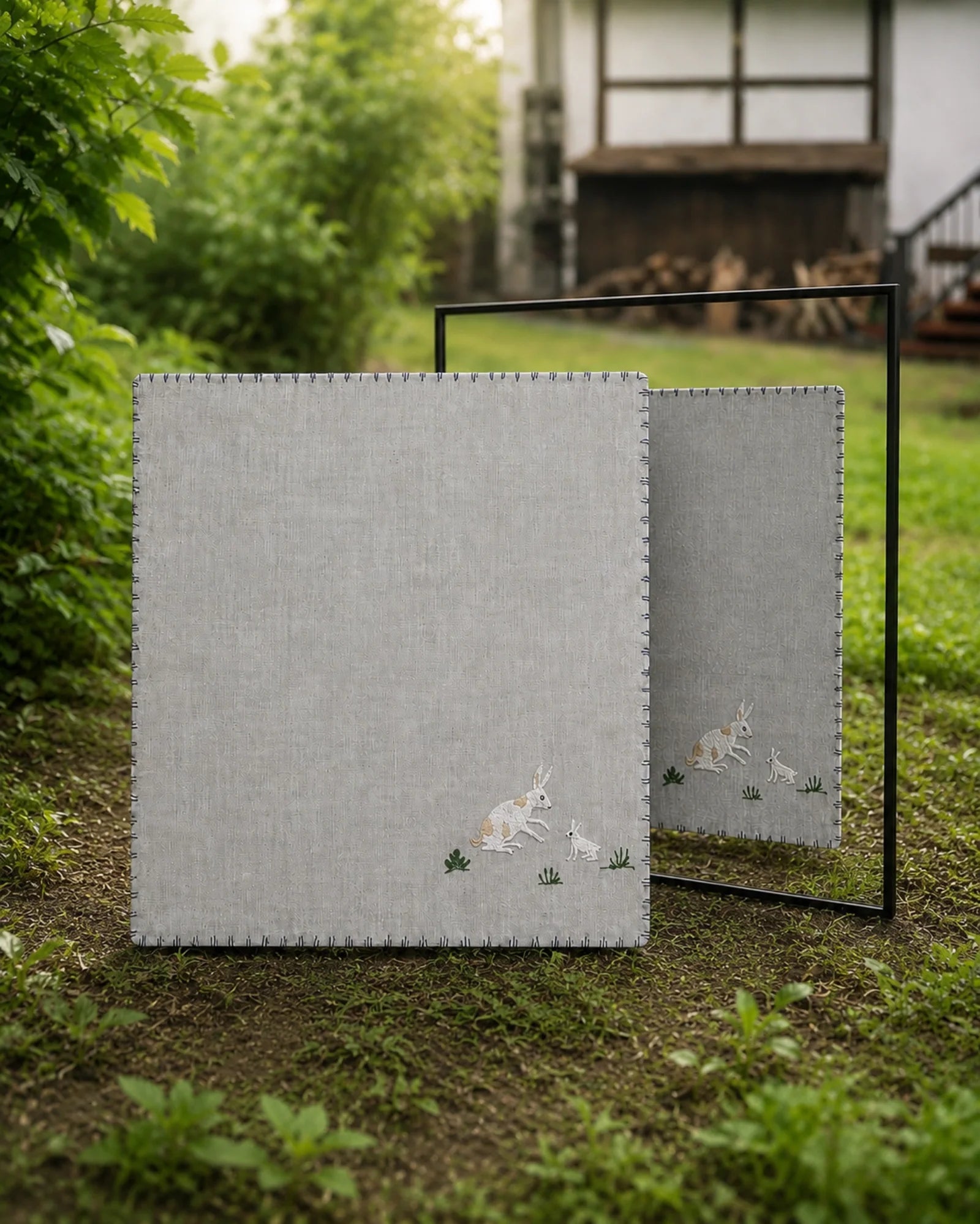 Chamba Rumal from the Jungle collection in handwoven khadi cotton, featuring a hand-embroidered rabbit and young rabbit among small grass, shown in a garden setting with a mirror revealing identical do-rukha embroidery on both sides.