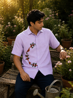Gond hand-painted linen shirt featuring a half-circle bird-and-tree motif composition across the right side of the shirt front, photographed while seated on a weathered garden bench and observing flowering plants within a sunlit garden setting surrounded by potted blooms and soft late-afternoon light.