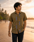 Gond hand-painted linen shirt featuring symmetrical botanical motifs flowing vertically along both sides of the button placket, photographed while walking barefoot along a quiet golden-hour shoreline with rolled black denim and soft coastal light.