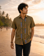 Gond hand-painted linen shirt featuring symmetrical botanical motifs flowing vertically along both sides of the button placket, photographed while walking barefoot along a quiet golden-hour shoreline with rolled black denim and soft coastal light.