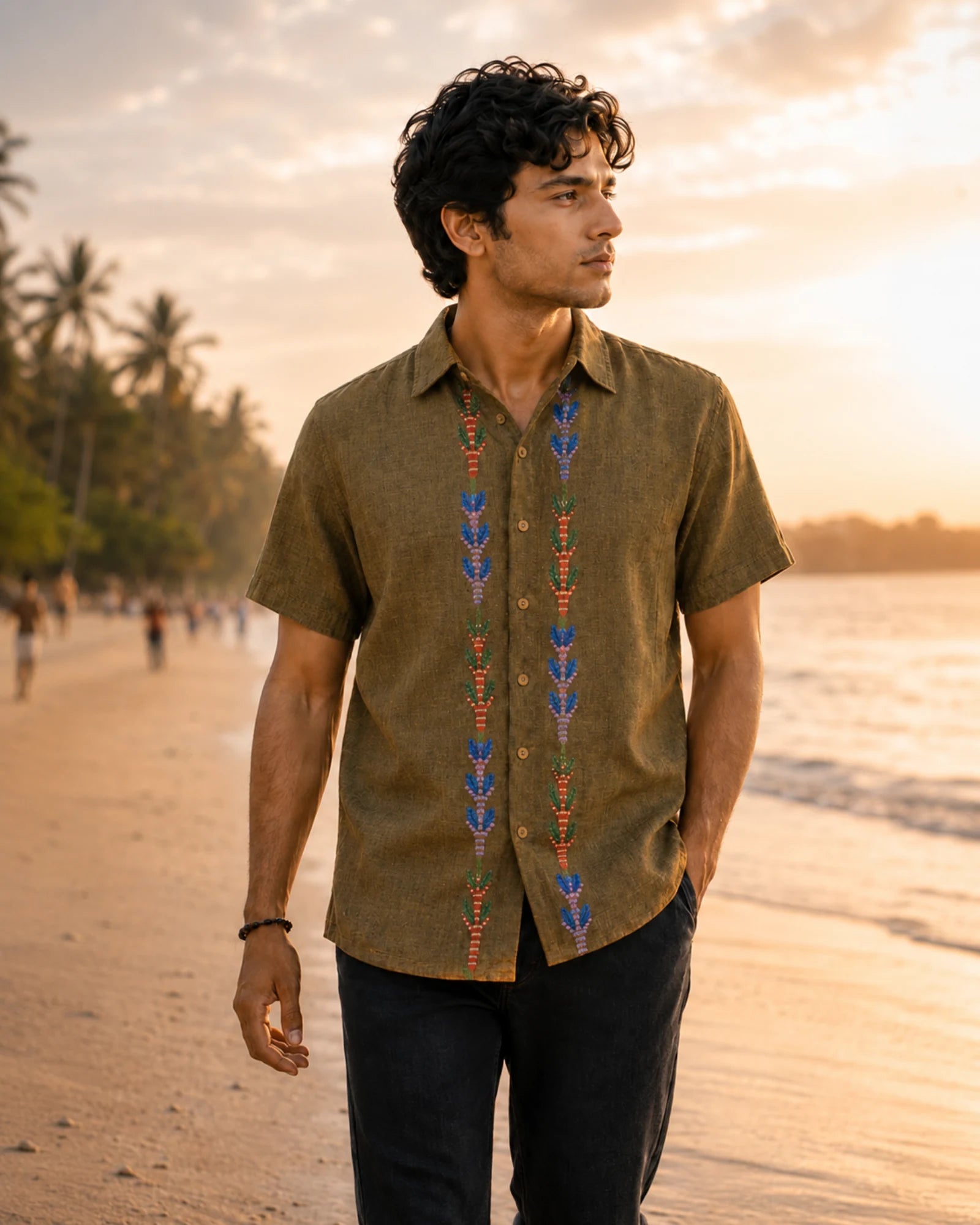 Gond hand-painted linen shirt featuring symmetrical botanical motifs flowing vertically along both sides of the button placket, photographed while walking barefoot along a quiet golden-hour shoreline with rolled black denim and soft coastal light.