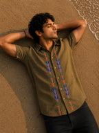 Gond hand-painted linen shirt featuring symmetrical botanical motifs flowing vertically along both sides of the button placket, observed from an overhead perspective while resting barefoot on warm shoreline sand beside gentle incoming waves within a quiet golden-hour coastal setting.