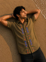 Load image into Gallery viewer, Gond hand-painted linen shirt featuring symmetrical botanical motifs flowing vertically along both sides of the button placket, observed from an overhead perspective while resting barefoot on warm shoreline sand beside gentle incoming waves within a quiet golden-hour coastal setting.