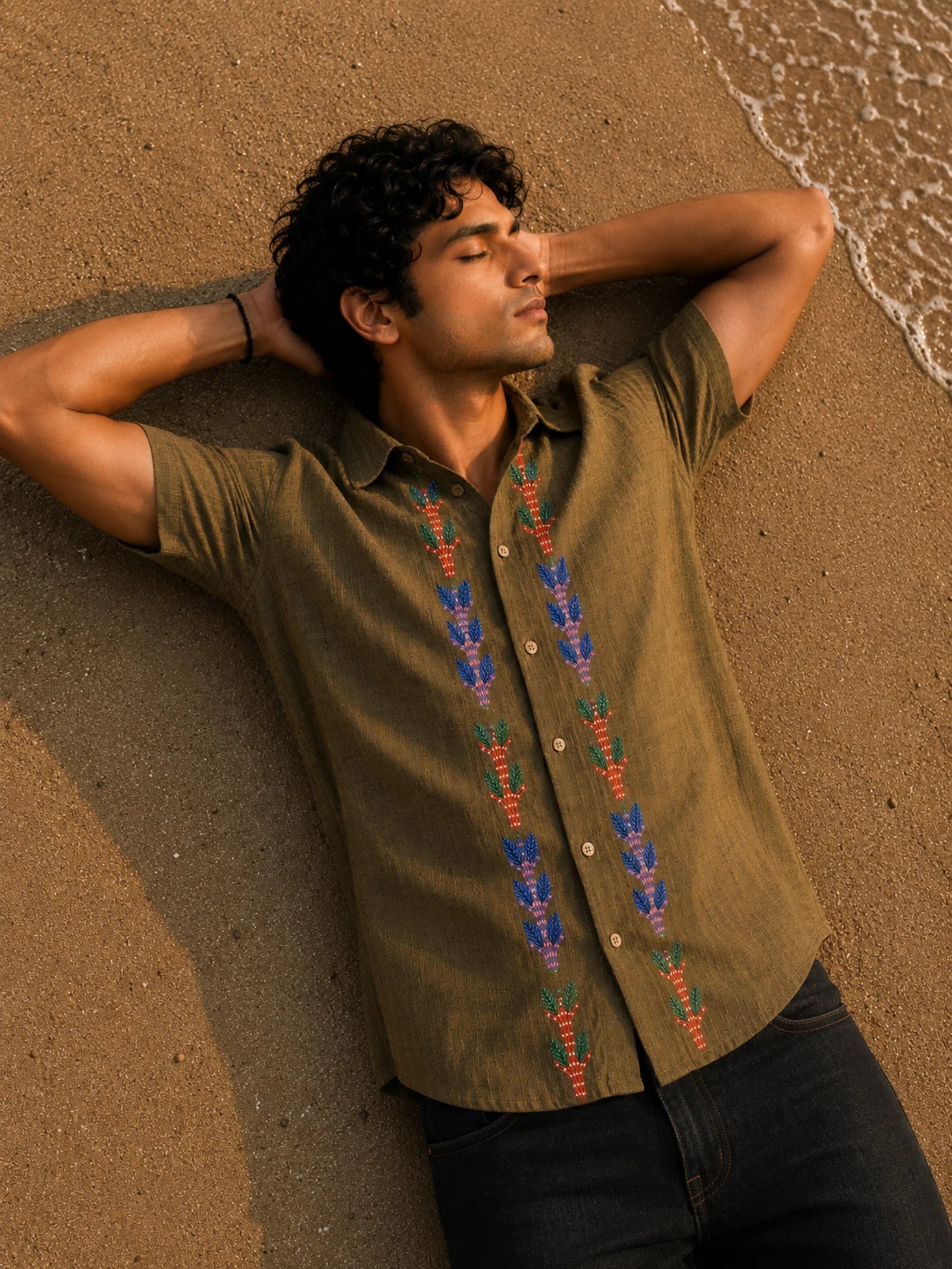 Gond hand-painted linen shirt featuring symmetrical botanical motifs flowing vertically along both sides of the button placket, observed from an overhead perspective while resting barefoot on warm shoreline sand beside gentle incoming waves within a quiet golden-hour coastal setting.