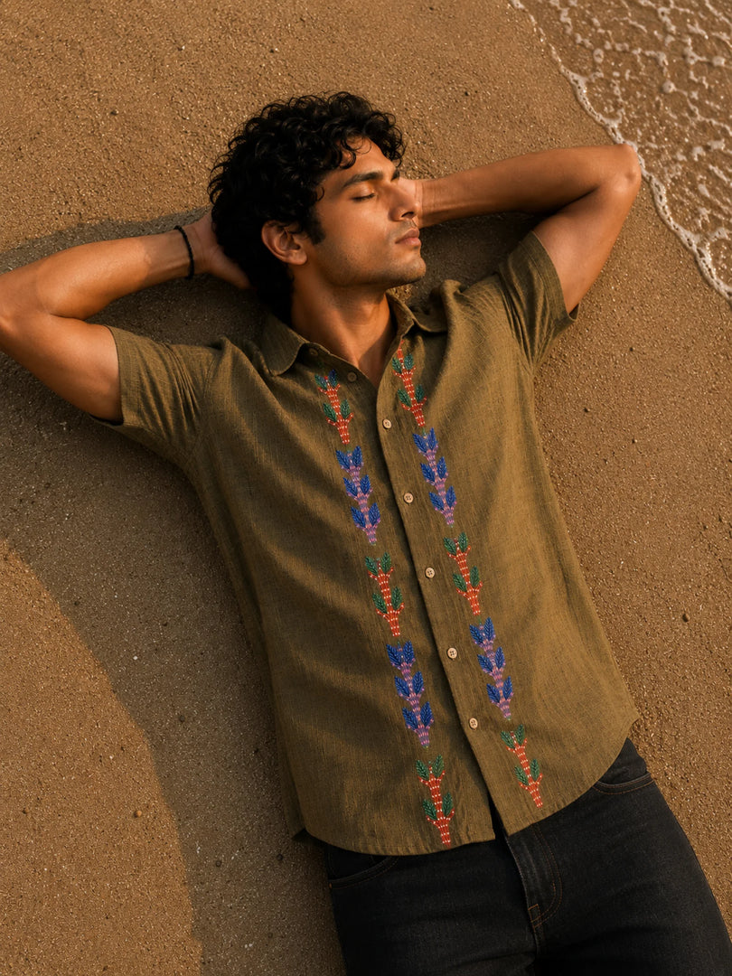 Gond hand-painted linen shirt featuring symmetrical botanical motifs flowing vertically along both sides of the button placket, observed from an overhead perspective while resting barefoot on warm shoreline sand beside gentle incoming waves within a quiet golden-hour coastal setting.