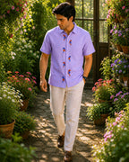 Gond hand-painted linen shirt featuring seven multicoloured bird motifs arranged vertically along the front placket, photographed while walking through a flowering garden pathway surrounded by potted blooms, lush foliage, and warm natural daylight.