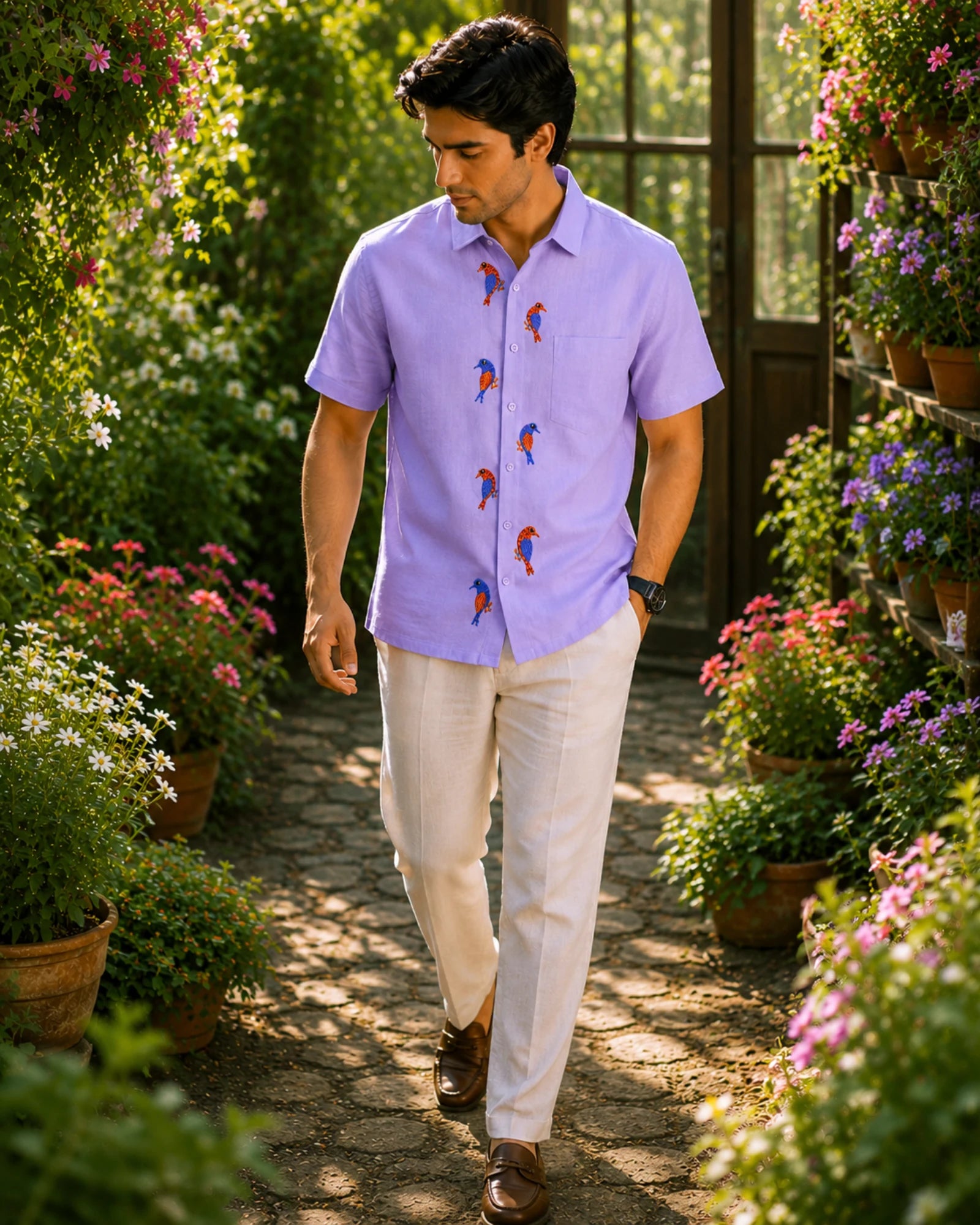 Gond hand-painted linen shirt featuring seven multicoloured bird motifs arranged vertically along the front placket, photographed while walking through a flowering garden pathway surrounded by potted blooms, lush foliage, and warm natural daylight.