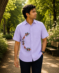 Gond hand-painted linen shirt featuring bird and branching tree motifs arranged in a half-circle composition flowing across the mid-right section of the shirt front, photographed in a quiet tree-lined garden pathway with black denim, soft natural daylight, and layered botanical surroundings creating a calm archival outdoor portrait atmosphere.