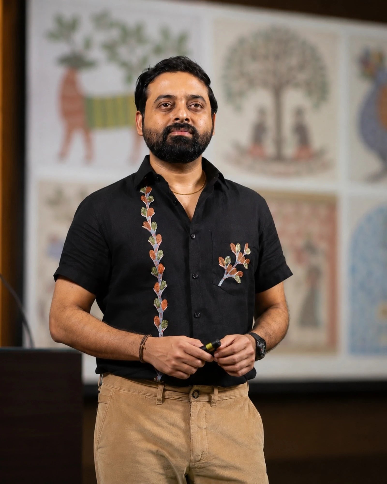 Gond hand-painted linen shirt featuring branching tree forms and leaf motifs extending across the placket and chest pocket, observed during an archival presentation setting before softly blurred folk art panels under warm interior lighting.