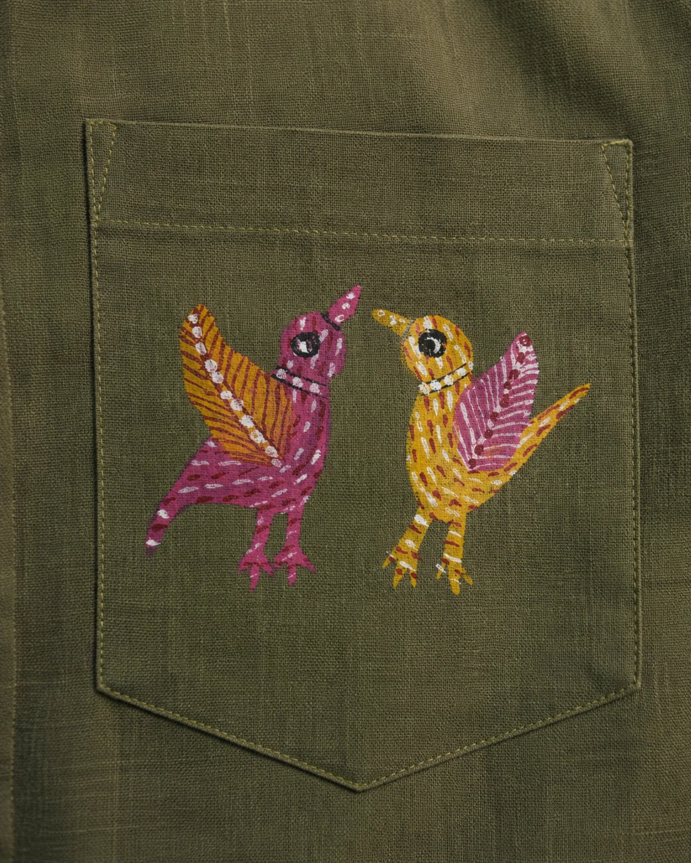 Close-up of a linen shirt pocket featuring two hand-painted Gond bird forms facing each other, constructed through layered linear brush detailing with visible pocket construction and textile weave across the linen surface.