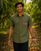 Gond hand-painted linen shirt featuring two bird forms positioned on the front pocket, photographed within a mango orchard setting while carrying a basket of raw and ripening mangoes in natural light.