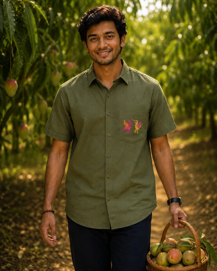Gond hand-painted linen shirt featuring two bird forms positioned on the front pocket, photographed within a mango orchard setting while carrying a basket of raw and ripening mangoes in natural light.