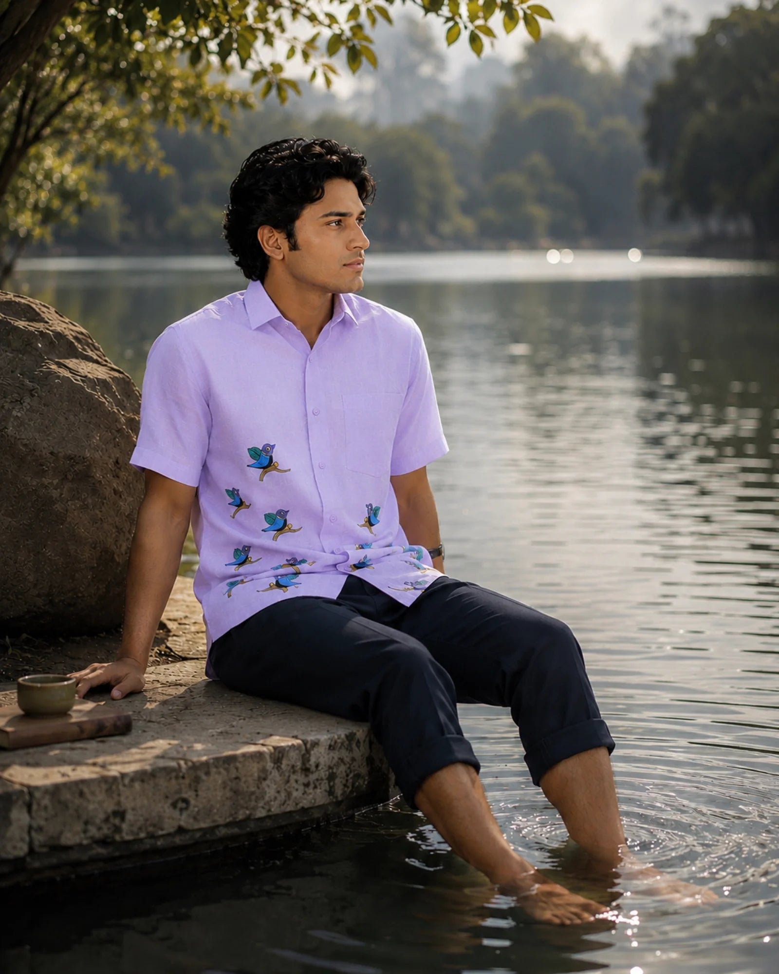 Gond hand-painted linen shirt featuring scattered bird motifs across the lower front panel, observed while seated beside a quiet lakeside edge with feet immersed in still water, reflecting within a contemplative natural setting under soft filtered daylight.