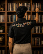 Gond hand-painted linen shirt featuring branching botanical forms extending across the upper back panel, observed in an archival library setting with softly blurred bookshelves and warm ambient lighting.