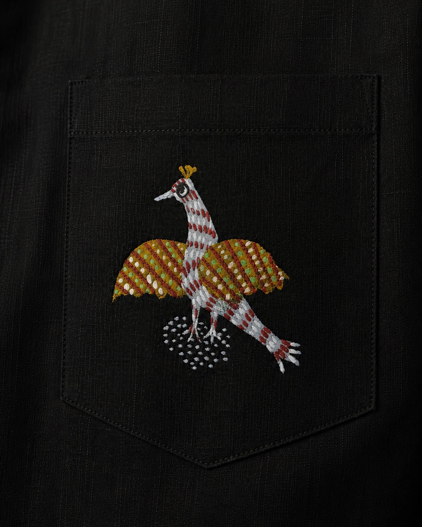 Close-up of a linen shirt pocket featuring a hand-painted Gond bird motif, revealing patterned linear detailing, pigment variation, pocket construction, and visible textile weave.
