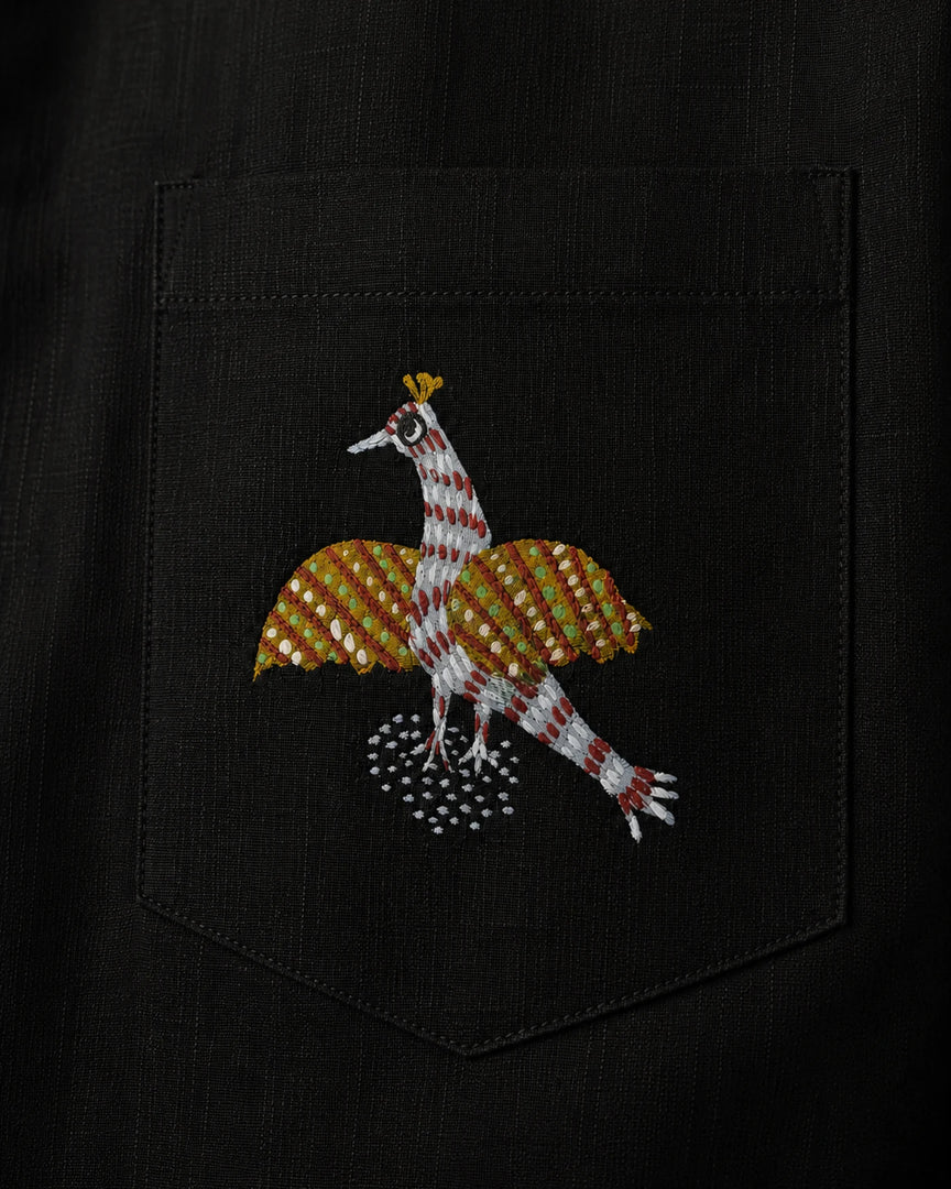 Close-up of a linen shirt pocket featuring a hand-painted Gond bird motif, revealing patterned linear detailing, pigment variation, pocket construction, and visible textile weave.