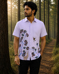 Gond hand-painted linen shirt featuring birds, branching tree forms, deer figures, and flowering elements flowing across the front panel, photographed while walking through a mist-covered forest trail in soft early morning light with tall woodland trees fading into the background.