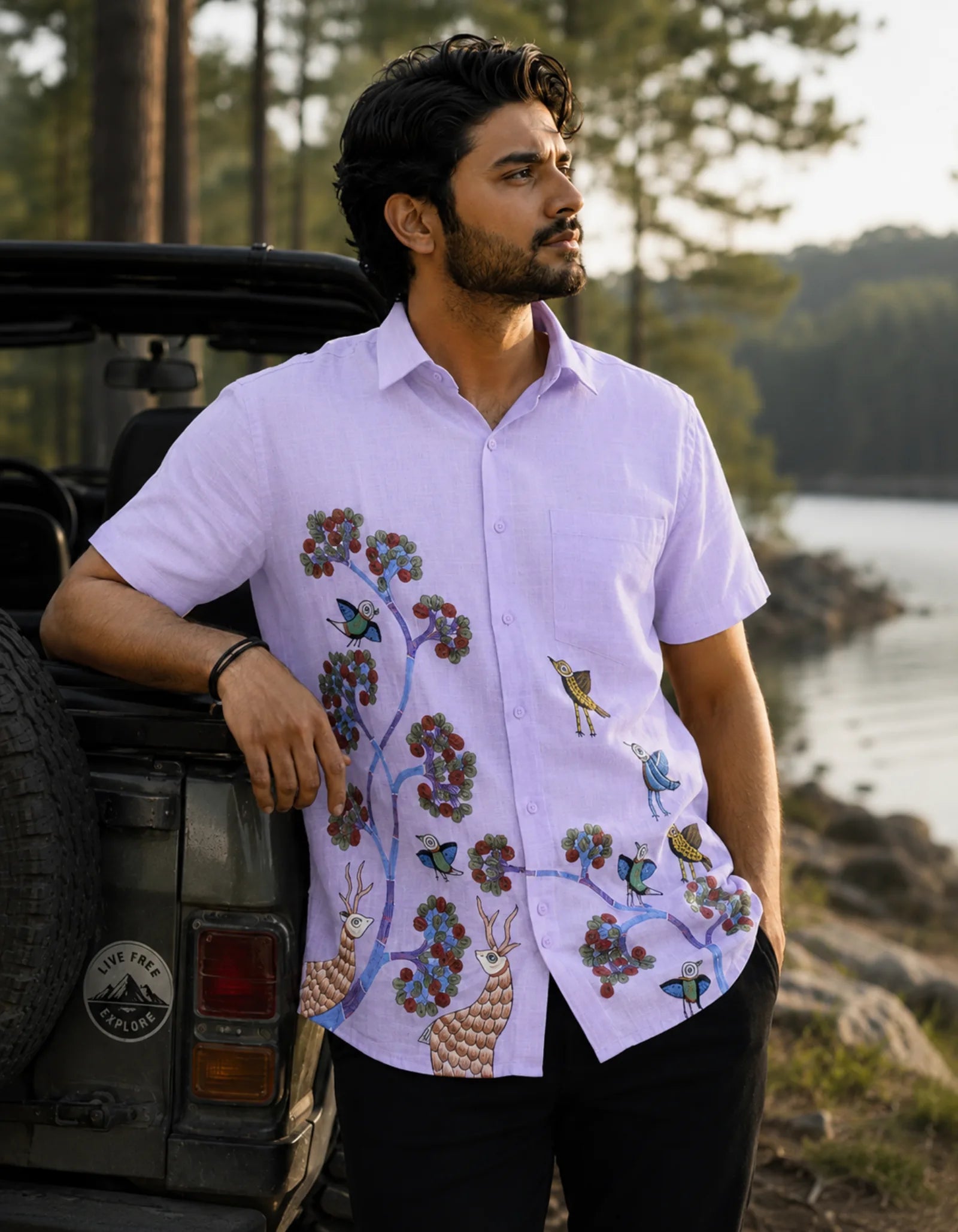 Gond hand-painted linen shirt featuring birds, branching tree forms, and deer motifs flowing across the front panel, observed while leaning against an open jeep beside a quiet lake and watching the surrounding trees within a soft golden-hour wilderness setting.