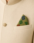 Alaap hand embroidered Kasuti pocket square crafted in 100% Mulmul Khadi, featuring peacock and geometric forms articulated through cobalt blue, emerald green and vermilion red threadwork. Resting within the pocket of an ivory ceremonial jacket, the composition draws attention to the flowing structure of counted Kasuti embroidery, the translucent softness of handwoven khadi, and the understated dialogue between Indian formalwear and living craft traditions under museum-toned illumination.