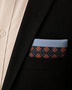 Arka Kasuti pocket square in 100% Mulmul Khadi featuring a Swastik motif rendered through counted hand embroidery in deep indigo and ember-toned threadwork. Styled within the breast pocket of a black tailored jacket, the image draws attention to the disciplined border geometry of Kasuti embroidery, the softened grain of handwoven Mulmul Khadi, and a restrained ceremonial character shaped through quiet archival composition and low, directional lighting.