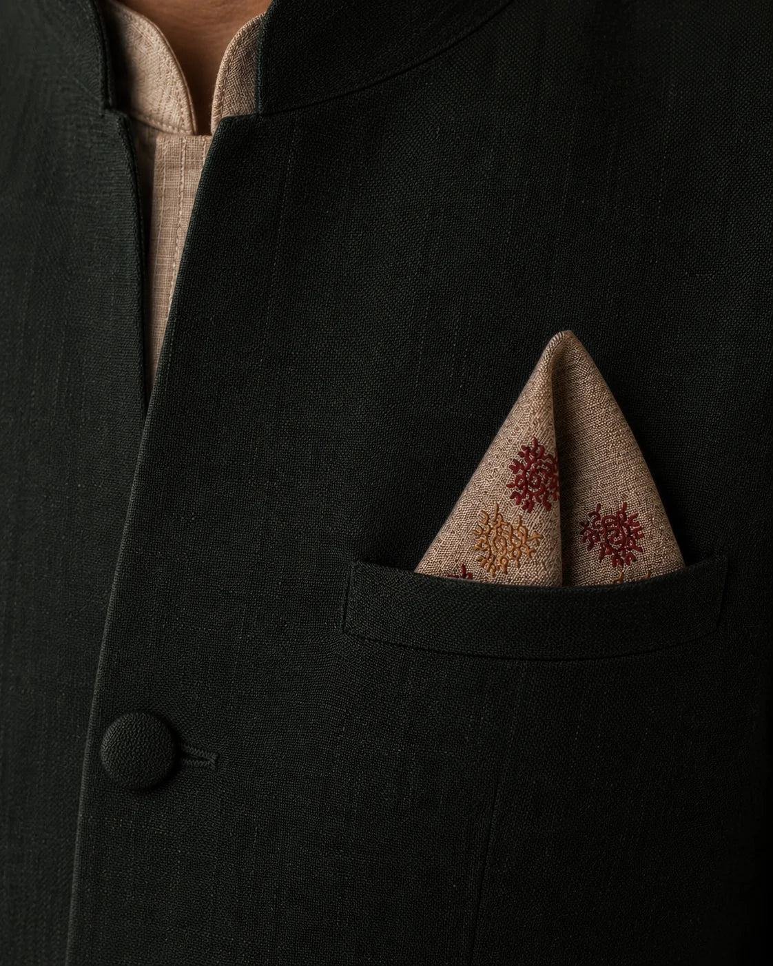 Dhvani hand embroidered Kasuti pocket square in 100% Mulmul Khadi featuring peacock and interconnected geometric motifs rendered in garnet red and burnished copper threadwork. Folded within the pocket of a deep forest-toned Nehru jacket, the composition brings together ceremonial Indian tailoring and the tonal rhythm of counted Kasuti embroidery, while the softened khadi weave and restrained archival lighting preserve a tactile sense of depth and enduring handcrafted character.