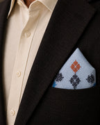 Laya Kasuti pocket square in 100% Mulmul Khadi featuring peacock and geometric motifs embroidered in deep indigo, softened sky blue, and muted coral threadwork. Styled within the breast pocket of a dark wood-toned tailored jacket against an ivory shirt, the image emphasizes the flowing cadence of counted Kasuti embroidery, the airy grain of handwoven Mulmul Khadi, and a composed archival mood shaped through restrained lighting and quiet formal dressing.
