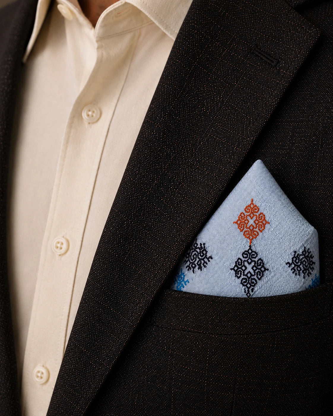 Laya Kasuti pocket square in 100% Mulmul Khadi featuring peacock and geometric motifs embroidered in deep indigo, softened sky blue, and muted coral threadwork. Styled within the breast pocket of a dark wood-toned tailored jacket against an ivory shirt, the image emphasizes the flowing cadence of counted Kasuti embroidery, the airy grain of handwoven Mulmul Khadi, and a composed archival mood shaped through restrained lighting and quiet formal dressing.