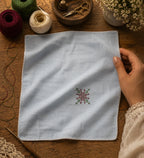 Kasuti hand embroidered pocket square on 100% Mulmul Khadi featuring a floral geometric motif in deep maroon and moss green threadwork, finished with hand-crocheted edging and photographed within a handcrafted textile workspace alongside embroidery threads, crochet lace, tailoring tools, wooden surfaces, and visible hand interaction that reflects the quiet patience and continuity of prolonged hand embroidery practice.