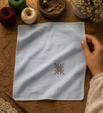 Load image into Gallery viewer, Kasuti hand embroidered pocket square on 100% Mulmul Khadi featuring a floral geometric motif in deep maroon and moss green threadwork, finished with hand-crocheted edging and photographed within a handcrafted textile workspace alongside embroidery threads, crochet lace, tailoring tools, wooden surfaces, and visible hand interaction that reflects the quiet patience and continuity of prolonged hand embroidery practice.