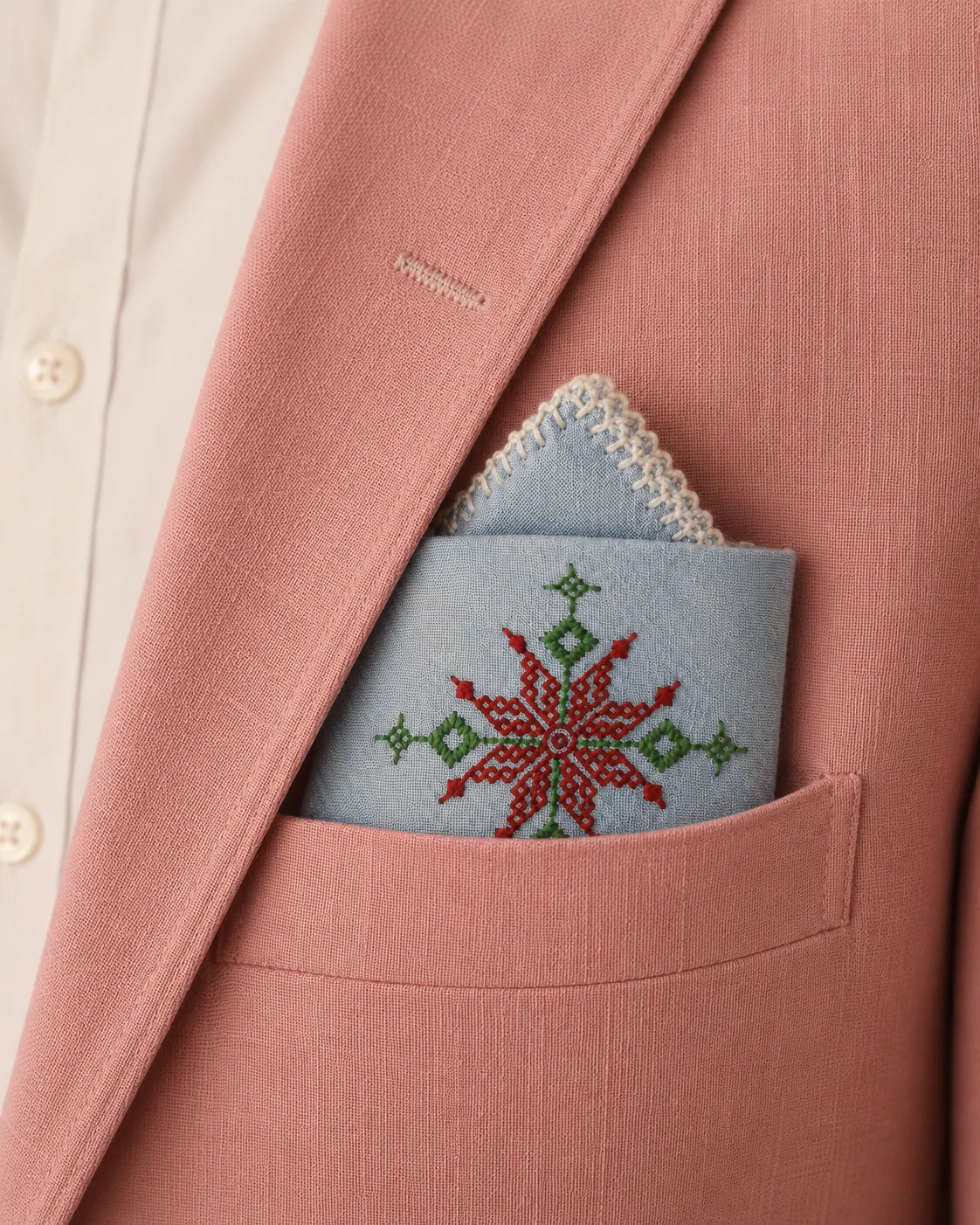 Hand embroidered Kasuti pocket square in 100% Mulmul Khadi featuring a lotus geometric motif rendered in deep maroon and moss green threadwork, folded within the breast pocket of a blush rose tailored jacket layered over an ivory shirt. The image balances the softened surface of Mulmul Khadi with the structured rhythm of counted Kasuti embroidery, creating a quiet dialogue between refined tailoring and hand-executed textile craft under restrained tonal lighting.
