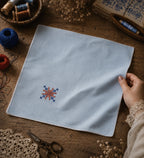 Kasuti hand embroidered pocket square on 100% Mulmul Khadi featuring a lotus geometric motif in ember and indigo threadwork, finished with hand-crocheted edging and photographed within a handcrafted textile workspace alongside embroidery threads, crochet lace, tailoring scissors, dried florals, wooden weaving tools, archival textile studies, and visible hand interaction that reflects the quiet continuity and patience of prolonged hand embroidery practice.