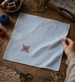 Load image into Gallery viewer, Kasuti hand embroidered pocket square on 100% Mulmul Khadi featuring a lotus geometric motif in ember and indigo threadwork, finished with hand-crocheted edging and photographed within a handcrafted textile workspace alongside embroidery threads, crochet lace, tailoring scissors, dried florals, wooden weaving tools, archival textile studies, and visible hand interaction that reflects the quiet continuity and patience of prolonged hand embroidery practice.
